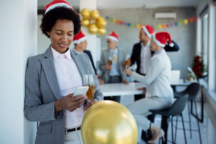 Happy black businesswoman using mobile phone and text messaging while having New Year's party in the office. Her colleagues are in the background.